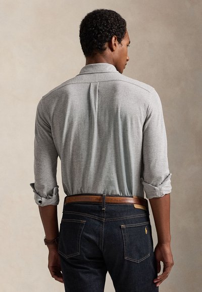 Mand med kort krøllet hår står med ryggen til iført en lysegrå langærmet skjorte med opkørte ærmer og mørkeblå jeans med et brunt bælte.