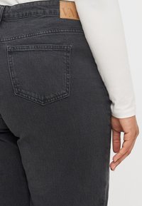 Selän puoli henkilöstä, joka pukeutuu tummanharmaisiin farkkuihin ja valkoiseen pitkähihaiseen paitan, näkyvillä takatasku ja osa kädestä, joka lepää sivulla.