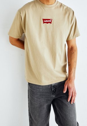 Beige bomulds T-shirt med korte ærmer, med et rødt broderet Levi's logo patch på forsiden; båret med grå denim jeans.