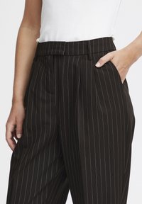 Zwarte broek met streepjes en plooien, met een tailleband en zijzakken. Stof lijkt glad met een subtiele glans.