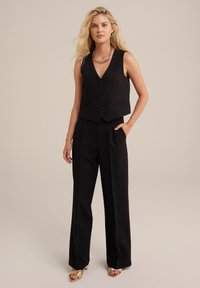 Črn skrajšan top z V-izrezom in dvema sprednjima žepoma, v kombinaciji z visokim črnimi hlačami in zlatimi sandalami, obogatena z verižnim ogrlicom.