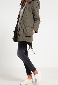 Olivově zelená parka bunda s kapucí vyloženou kožešinou, dvěma předními kapsami a stahováním v pase. Kombinována s tmavými džínami a bílými teniskami.