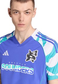 Ung mand iført en blå og teal Seattle Sounders fodboldsuniform med en logo af en spekkhugger og en fodbold på brystet, ser fremad med et alvorligt udtryk.