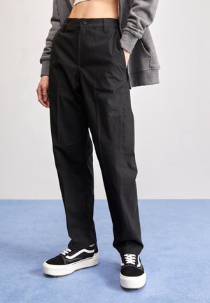 Osoba nosząca czarne spodnie o prostych nogawkach z logo The North Face, czarno-białe sneakersy oraz szarą bluzę z kapturem i zamkiem, z rękami w kieszeniach.