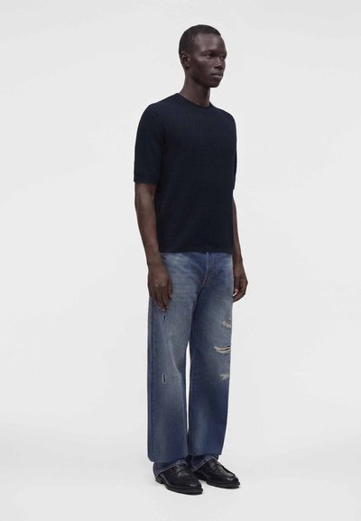 Homme debout portant une chemise noire à manches courtes, un jean bleu usé et des chaussures habillées noires sur un fond clair uni.