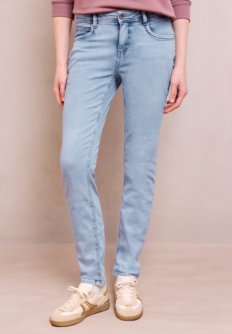 Personne portant un jean slim bleu clair, des baskets beige et blanches, et un haut à manches longues rose, debout devant un fond uni.