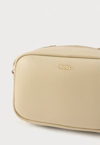 Mala de pele bege com forma rectangular, apresentando uma textura suave e um logótipo dourado "BOSS". Tem um fecho de correr na parte superior.