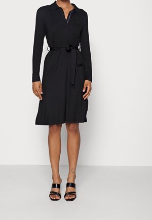 Woman wearing a knee-length long-sleeve black dress with collar, waist tie, and black open-toe heels standing against a white background.