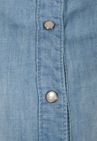 Gros plan sur un tissu bleu clair avec deux boutons-pression argentés sur une patte cousue.