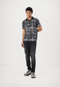 Polo de manga corta con un patrón geométrico en blanco y negro, con un cuello negro sólido. Combinado con jeans oscuros y zapatillas deportivas grises.
