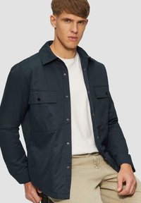 Granatowa koszula zapinana na guziki z kołnierzykiem, dwiema kieszeniami na piersiach i miękkiego materiału; noszona na białym T-shircie, w zestawie z beżowymi spodniami.