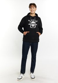 Sort hætte sweatshirt med hvid kraniumgrafik og guitar designs, med frontlommer og snørelukning. Båret med mørke denim jeans.