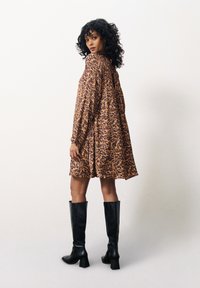 Braunes Leopardenmuster-Kleid mit langen Ärmeln und lockerer Passform, kombiniert mit hohen schwarzen Lederstiefeln mit Absatz. Glatte Textur und lässiges Design.