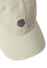 Gorra de algodón beige claro con una superficie texturizada, que presenta un parche de logo circular en blanco y negro en la parte frontal. Diseño de visera curvada.