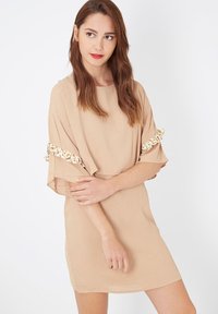 Joven mujer con cabello castaño claro viste un vestido beige con bordes florales en las mangas acampanadas, de pie frente a un fondo blanco liso.