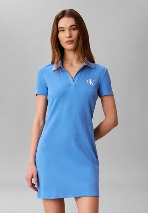 Kobieta w niebieskiej sukience polo z krótkim rękawem z logo Calvin Klein na piersi, stojąca na tle jednolitego szarego tła.