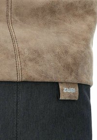 Bolso de cuero marrón con una superficie suave y texturizada y costuras visibles. Incluye una etiqueta de tela con el nombre de la marca, ubicada en un cuerpo de tela oscura.