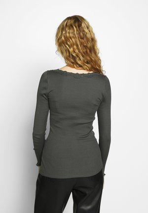 Kvinde med bølget blondt hår iført en tætsiddende mørkegrå ribstrikket langærmet top med blonderkant, set bagfra mod en hvid baggrund.