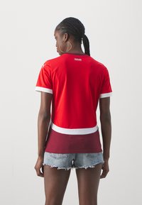 Sarkans sportisks T-krekls ar īsām piedurknēm un baltu apdari, ar logotipa uzlīmi aizmugurē un melnā sarkanā gradienta apakšmalu, saskaņots ar džinsu šortiem.