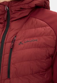 Giacca isolante rossa con design trapuntato. Presenta una tasca con zip e dettagli in zip neri, che mostrano il logo VAUDE in nero.