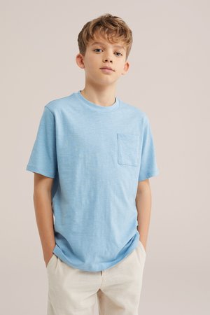 Chłopiec z jasnobrązowymi włosami, ubrany w jasnoniebieską koszulkę z krótkim rękawem z kieszenią na piersi i beżowe spodnie, stoi z rękami w kieszeniach.