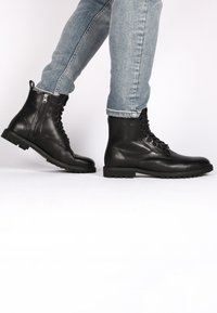 Botas de cuero negras hasta el tobillo con puntera redonda, diseño con cordones, cierre de cremallera lateral y suela de goma resistente, combinadas con jeans de color azul claro.