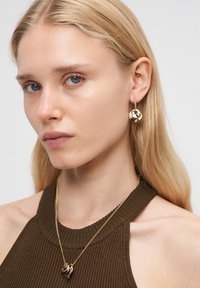 En blond kvinde med blå øjne bærer guld abstrakte øreringe, en guldpandant halskæde og en brun ærmeløs ribbet top foran en hvid baggrund.