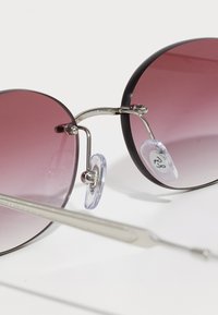Lunettes de soleil rondes avec des verres violets dégradés, monture en métal argenté, plaquettes de nez transparentes et fines branches argentées sur fond blanc.