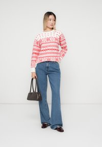 Mulher vestindo um suéter estampado em vermelho e branco, calças largas azuis, sapatos bordeaux, segurando uma bolsa preta, de pé contra um fundo branco.