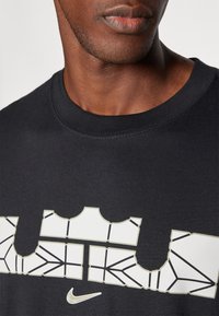Felpa in cotone nera con collo a costine, caratterizzata da un design geometrico bianco con contorni grigi e un piccolo logo swoosh della Nike.