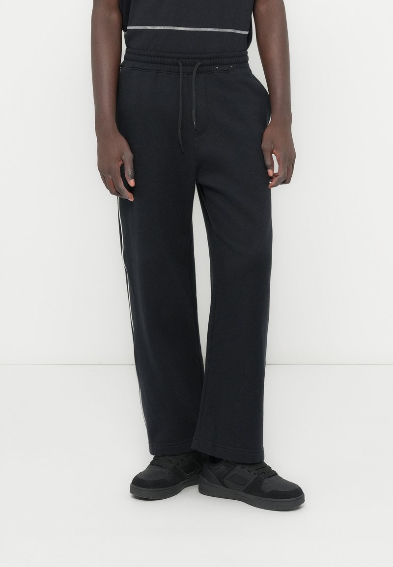 Μαύροι αθλητικοί παντελόνια με ελαστική μέση, πλαϊνές τσέπες με σχισμές και λευκές πλαϊνές ρίγες. Χαλαρή εφαρμογή και μαλακό ύφασμα, συνδυασμένα με μαύρα αθλητικά παπούτσια.