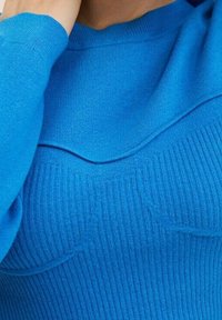 Pull tricoté bleu avec texture côtelée, présentant un col rond et des détails subtils de contour de poitrine. Manches et finition du tissu lisses.