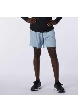 Persona in piedi con le mani sui fianchi che indossa pantaloncini sportivi azzurri, top a maniche lunghe nero e scarpe da corsa nere su sfondo semplice.