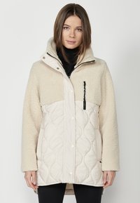 Beige prešívaná bunda s nadýchaným vrchným dielom pripomínajúcim vlnu, vysokým golierom, predným zipsom a patentkami, a vreckom na zips na hrudi.