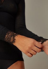 Schwarzes langärmliges Oberteil mit Spitze an den Handgelenken. Der Stoff scheint weich mit einer glatten Textur zu sein und verfügt über zarte Spitzenverzierungen.