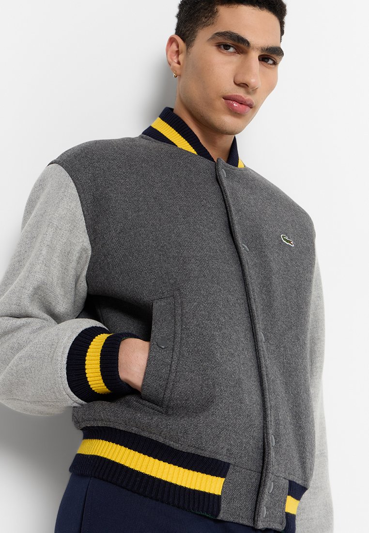 Veste en laine grise et gris clair avec un col côtelé bleu marine et jaune. Présente des boutons-pression et des poches latérales. Logo Lacoste sur la poitrine.