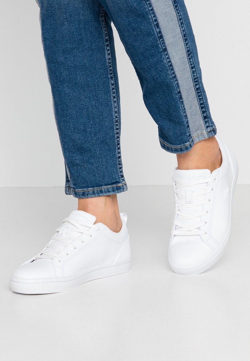 Personne portant un jean bleu avec des rayures latérales claires et des baskets blanches basses debout sur un sol gris.