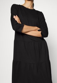 Femme portant une robe noire à manches longues à volants, debout, les bras croisés, devant un fond blanc uni.