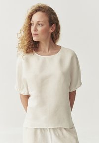 Light beige short-sleeve blouse with a round neckline and gathered sleeves, made from a textured, slightly sheer fabric.