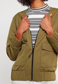 Olivgrön zip-jacka med ribbad textur, sidofickor och elastiska ärmslut, lager på lager över en svart- och vitrandig topp.