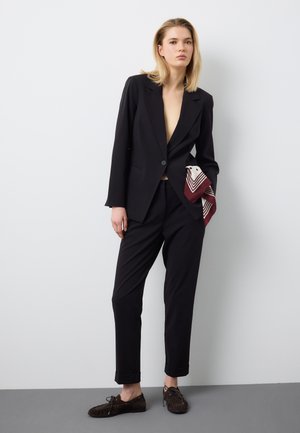 Donna con abito nero e blazer sbottonato che tiene una sciarpa rossa e bianca piegata, in piedi contro una parete bianca e un pavimento grigio.