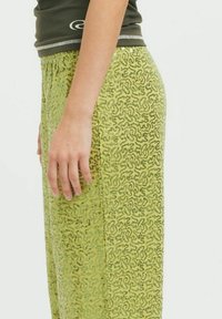 Zelene sekvinske hlače s teksturiranim vzorcem, širšega kroja in elastičnim pasom. Na strani je vidna roka.