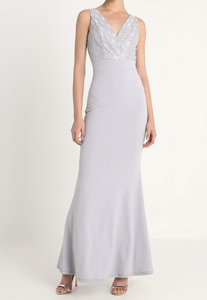 Mouwloze lichtgrijze avondjurk met een lijfje van pailletten en een V-hals, een aangesloten taille en een lange vloeiende rok, gedragen met zilveren open-toe hakken.