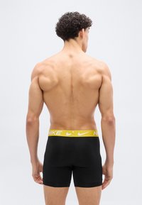 Μυώδης άνδρας με σγουρά μαλλιά, γυρισμένος πλάτη, φορώντας μαύρα μπόξερ της Nike με κίτρινη μέση.