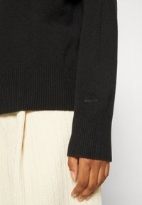 Gros plan d'une main portant une manche de pull côtelée noire sur une jupe crème texturée, avec un logo GANT subtil sur la manche.