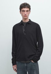Młody mężczyzna z krótkimi, kręconymi włosami, ubrany w czarną, długą koszulę z guzikami na jasnoszarym t-shircie i wyblakłych niebieskich dżinsach, stojący na jednolitym tle.