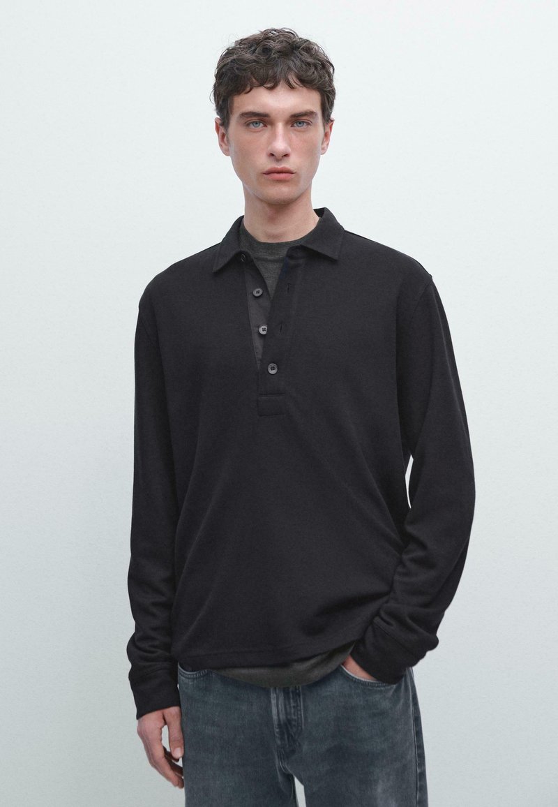 Ung man med kort lockigt hår, iklädd en svart långärmad skjorta med knappar över en mörkgrå t-shirt och blekta blåjeans, stående mot en enfärgad bakgrund.