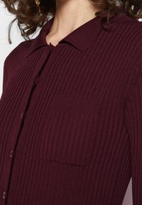 Cardigan bordeaux a coste con colletto, dotato di chiusura a bottoni e piccola tasca sul petto. Tessuto strutturato e righe verticali.