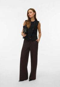 Svart ärmlös topp med strukturerad design och två knappar, kombinerad med vida mörkbruna byxor och håller i en svart clutchväska.