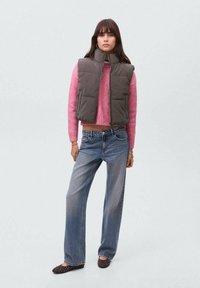 Gilet grigio scuro imbottito sopra un maglione lavorato a maglia rosa, abbinato a jeans blu larghi e ballerine marroni a pois. Outfit casual semplice.